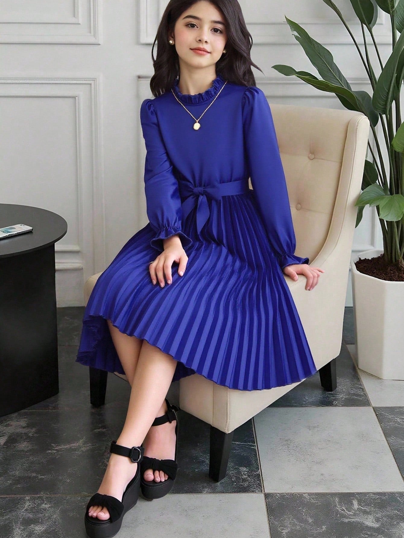 Tween girl wearing a royal blue long sleeve pleated casual A-line dress featuring a stand collar, flare sleeves, high waist with front tie belt, and ruffle hem.