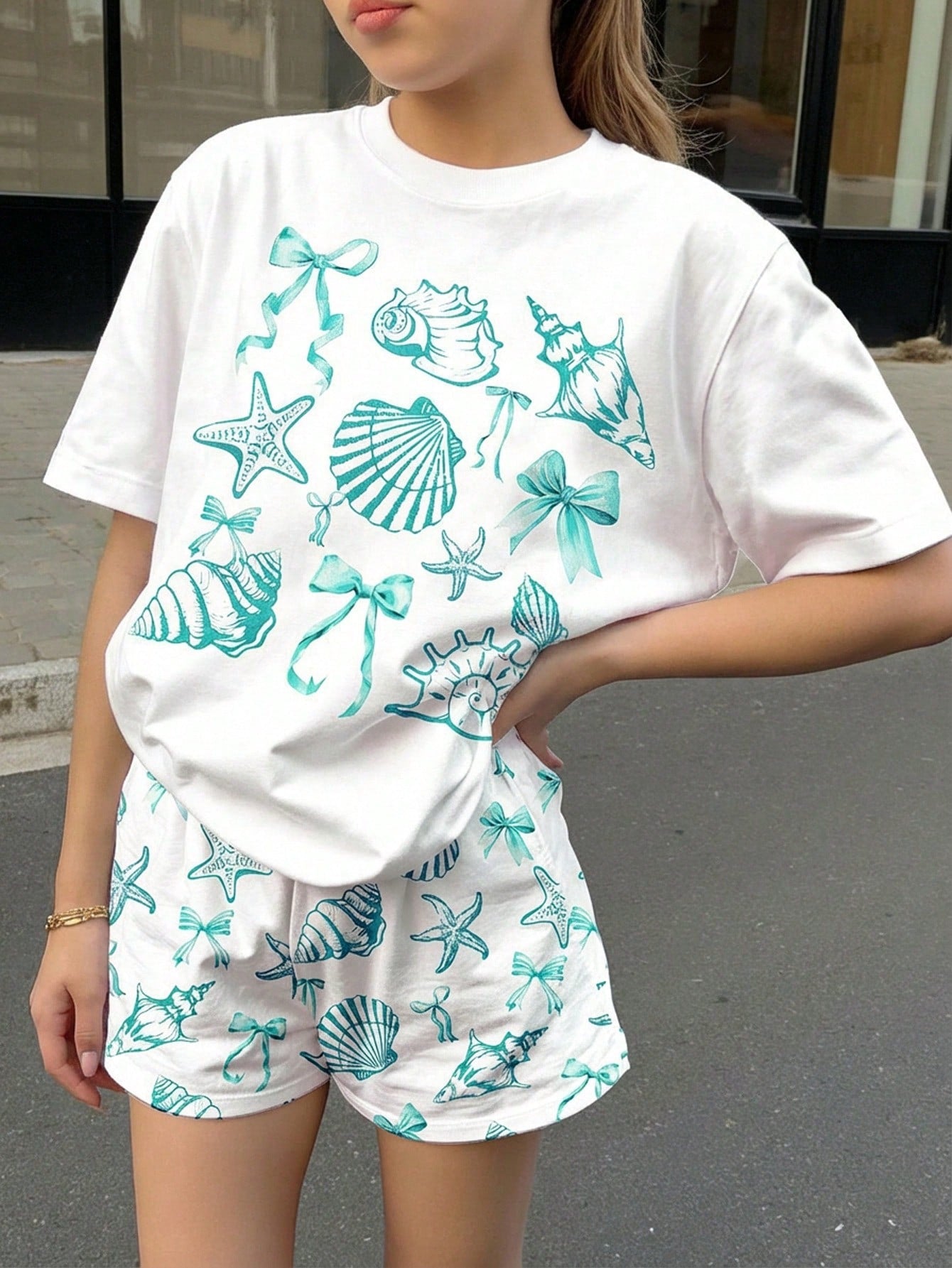 Teen girl wearing 2-piece summer vacation set with blue and white striped short sleeve top featuring seashell ocean bow print and matching loose fit shorts with pockets.