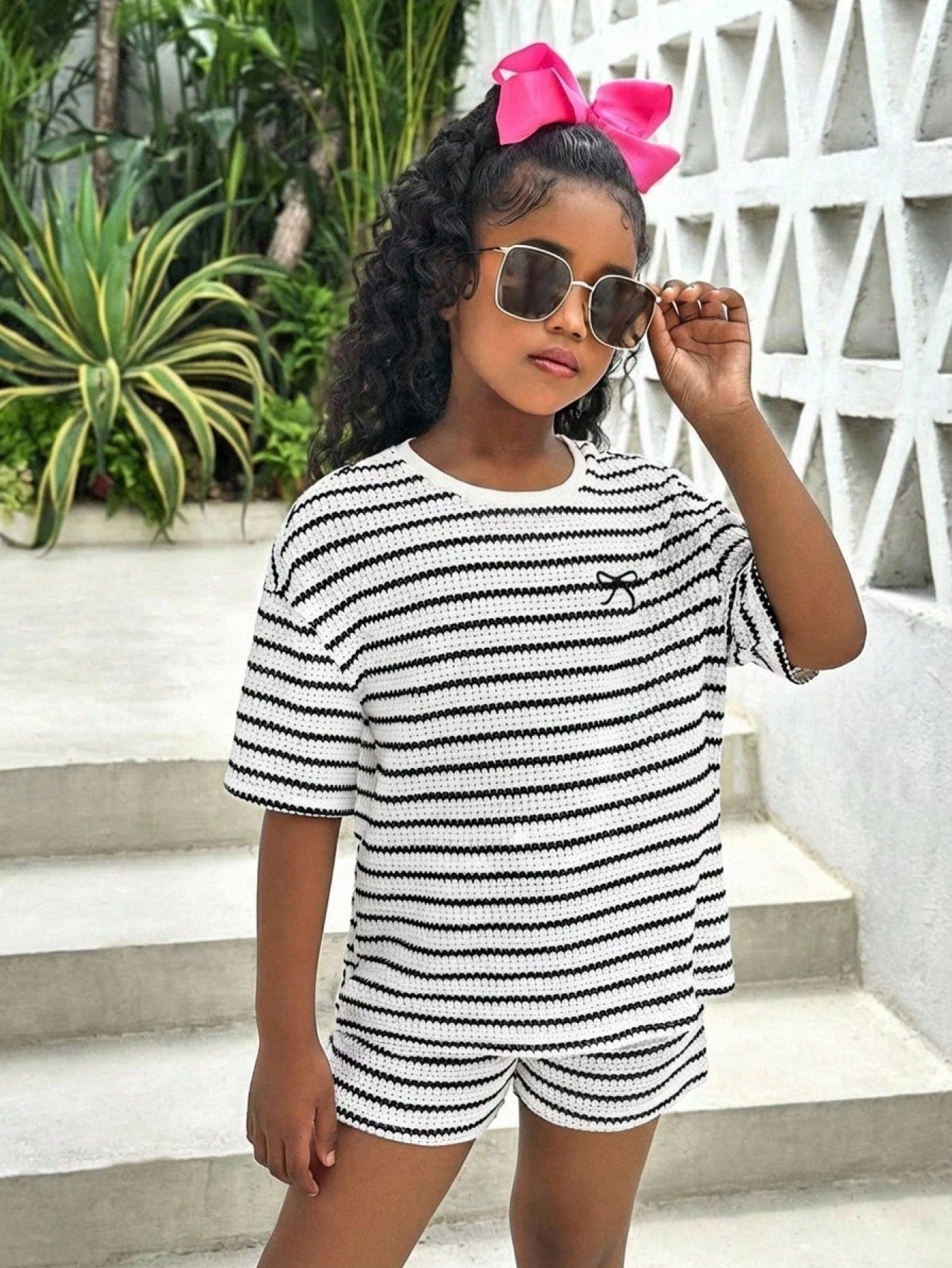 Tween girl wearing a black and white striped drop-shoulder crew neck t-shirt with bow embroidery paired with matching tie-waist shorts.