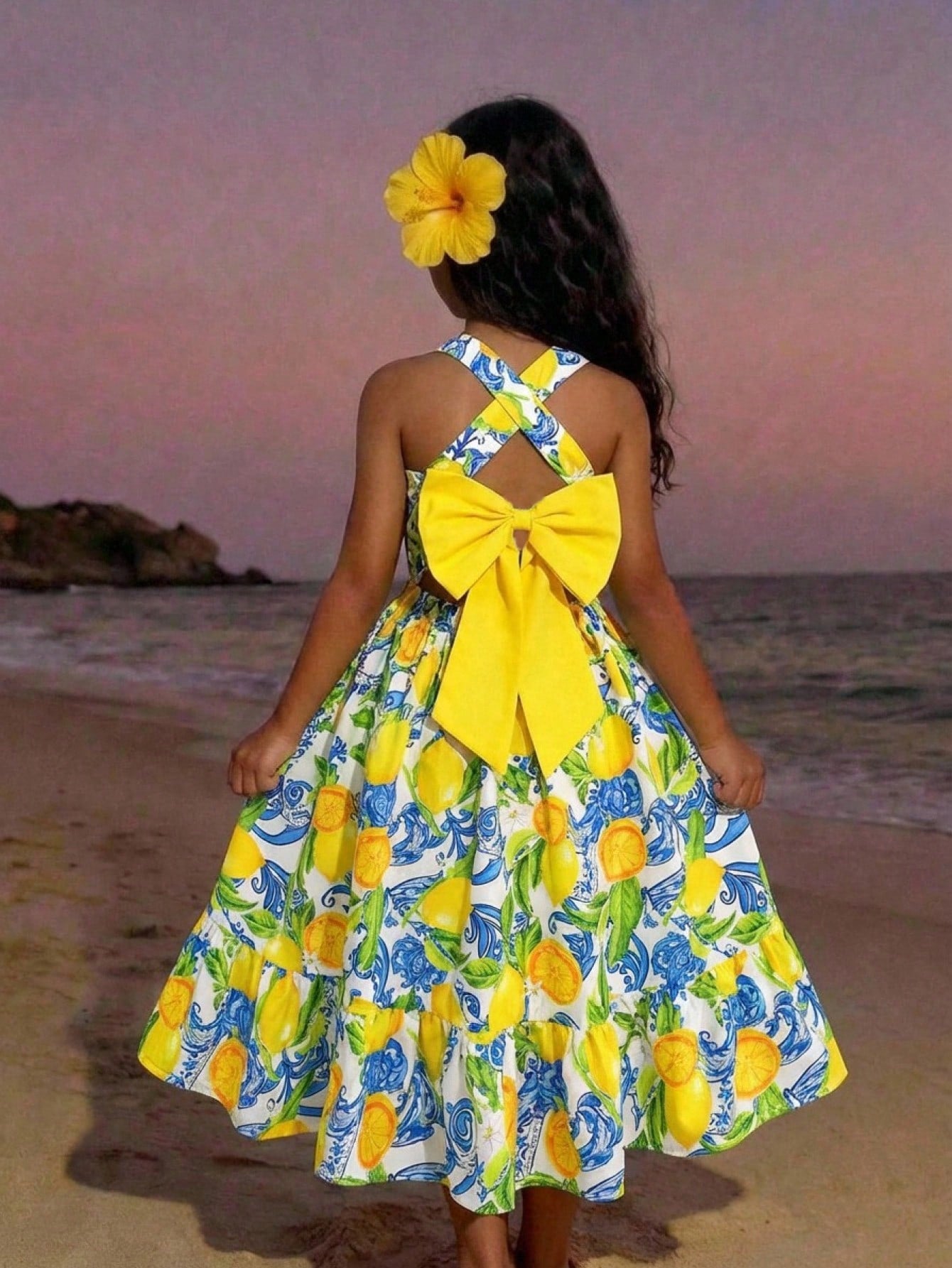 Pink sleeveless tween girls vacation dress with all-over fruit and vegetable print, strap neckline, belted high waist, cut-out detail, and flared ruffle hem.