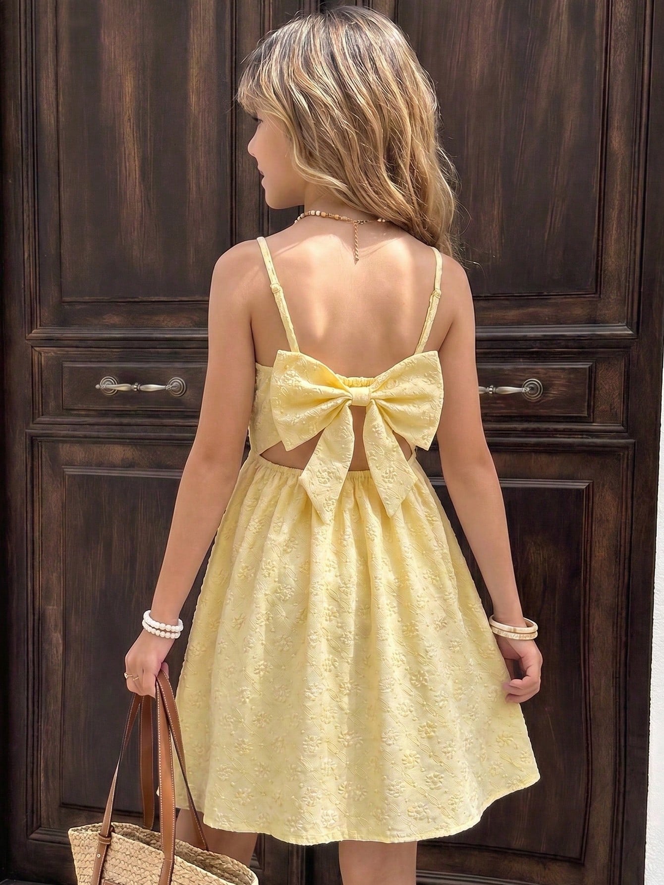Tween girl wearing an apricot floral print sleeveless mini cami dress featuring spaghetti straps, a shirred high waist, flounce hem, and a bow back detail.