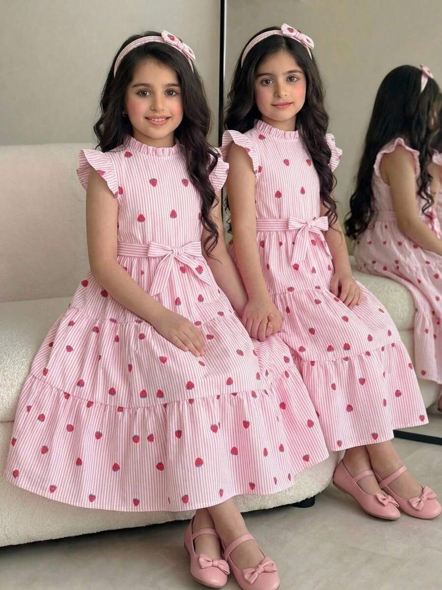Tween girl wearing a pink long A-line dress featuring a strawberry print, stand collar, flounce short sleeves, and a belted waist with a ruffle hem.