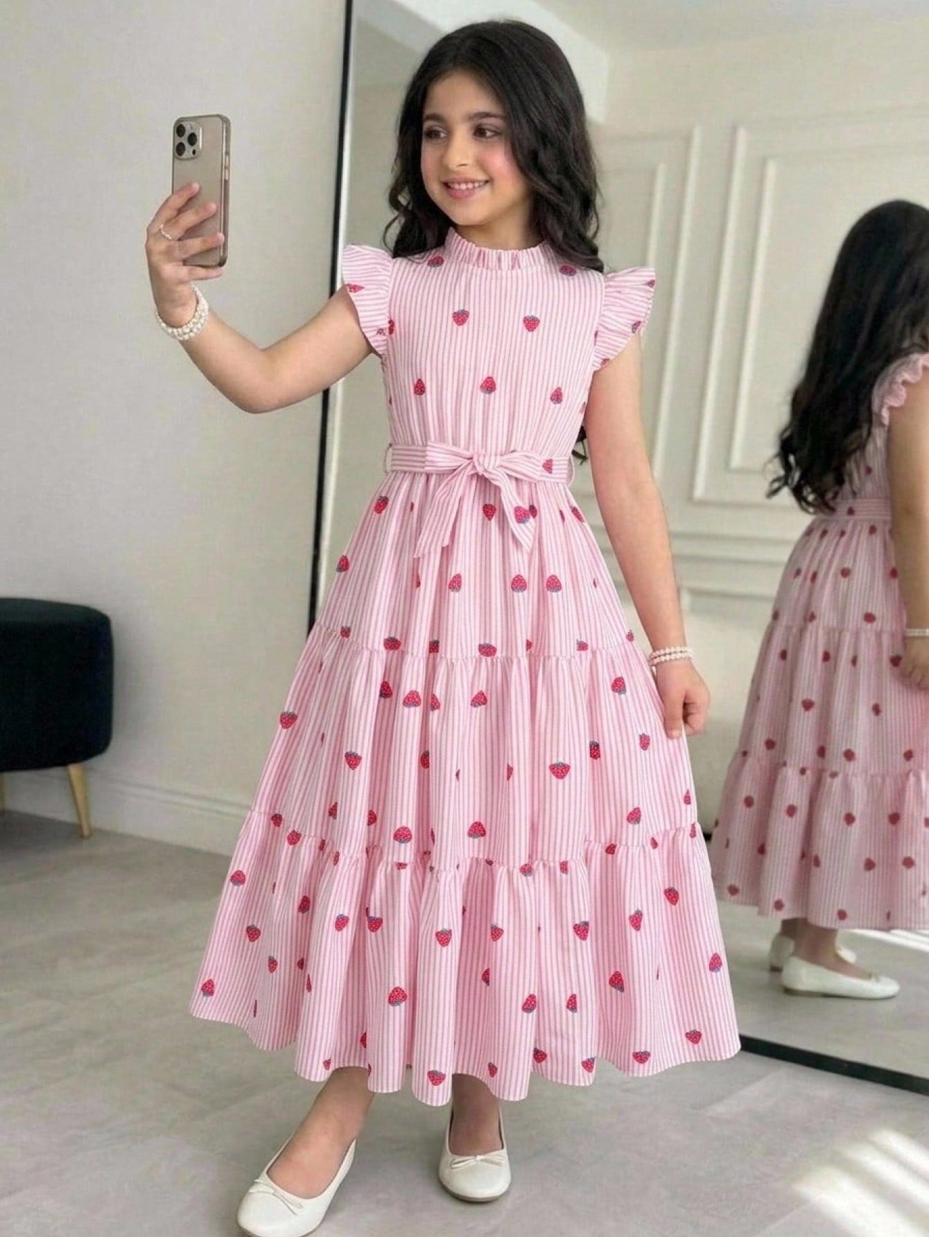 Tween girl wearing a pink long A-line dress featuring a strawberry print, stand collar, flounce short sleeves, and a belted waist with a ruffle hem.