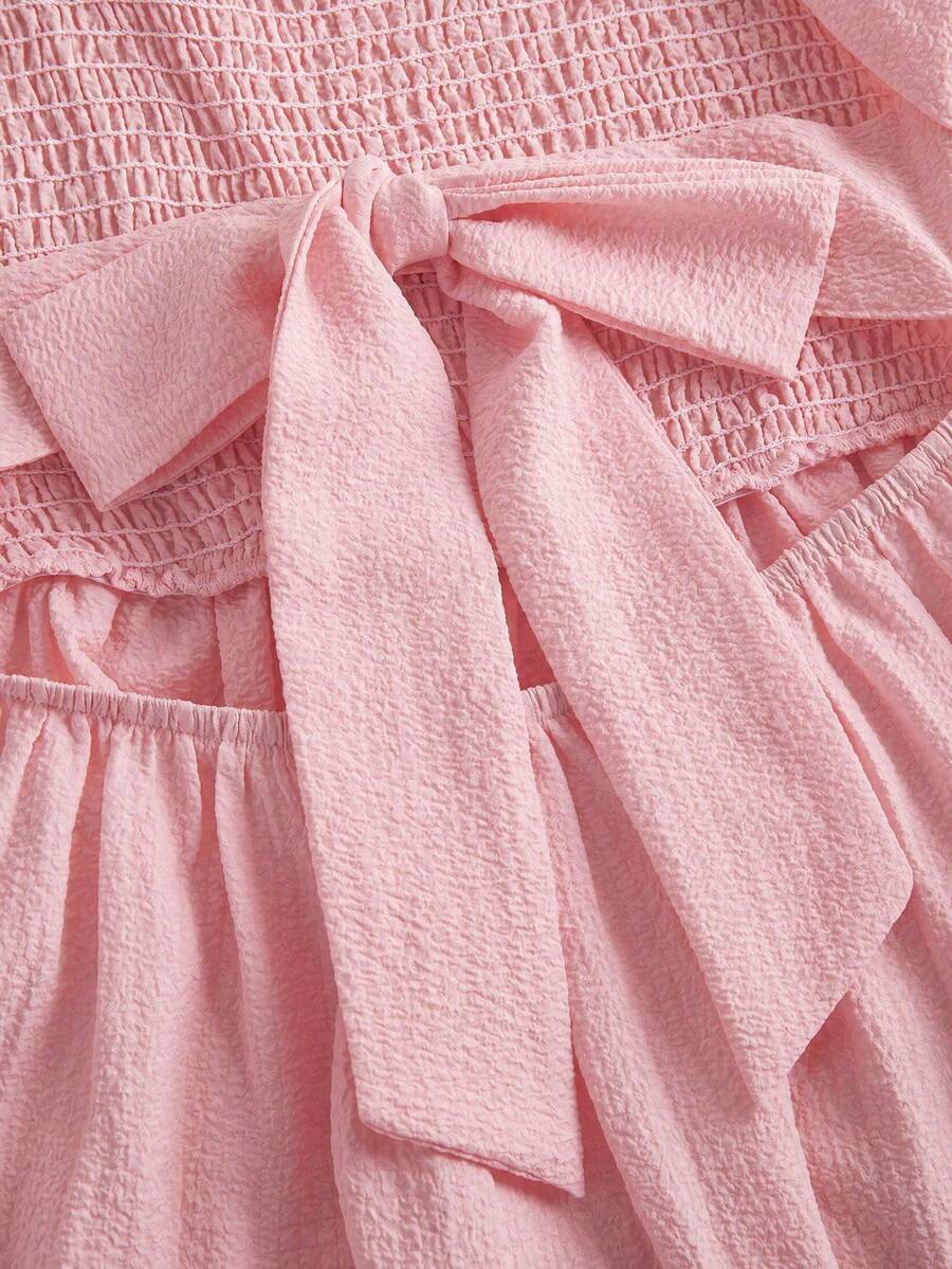 Pink sleeveless tween girls vacation dress with all-over fruit and vegetable print, strap neckline, belted high waist, cut-out detail, and flared ruffle hem.