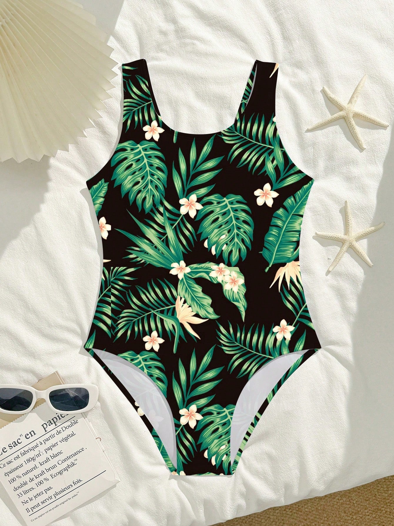 Tween girls one-piece swimsuit with a black base and multicolor tropical plant print featuring bamboo, palm leaves, plumeria flowers, scoop neck, and wide straps.