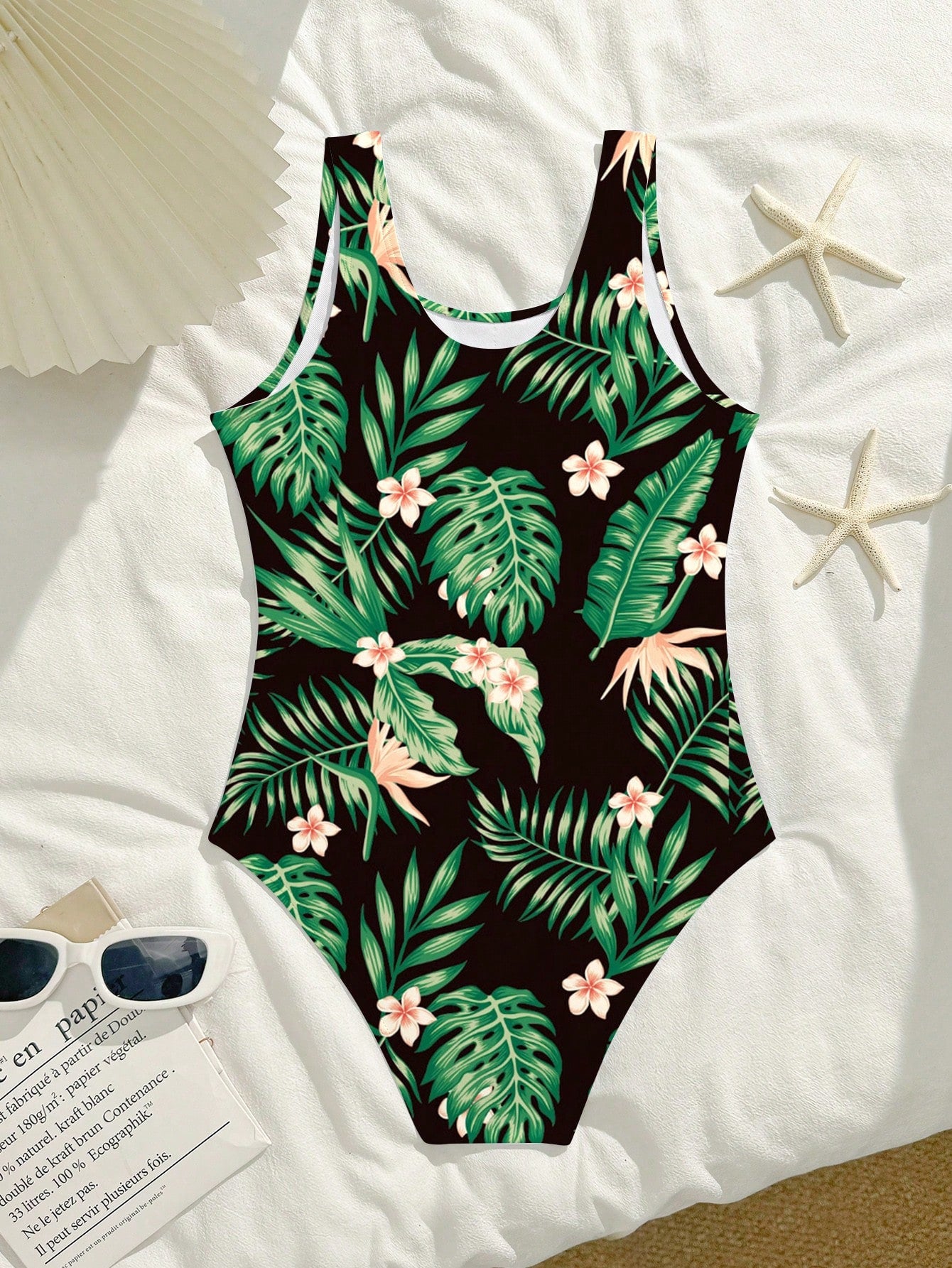 Tween girls one-piece swimsuit with a black base and multicolor tropical plant print featuring bamboo, palm leaves, plumeria flowers, scoop neck, and wide straps.
