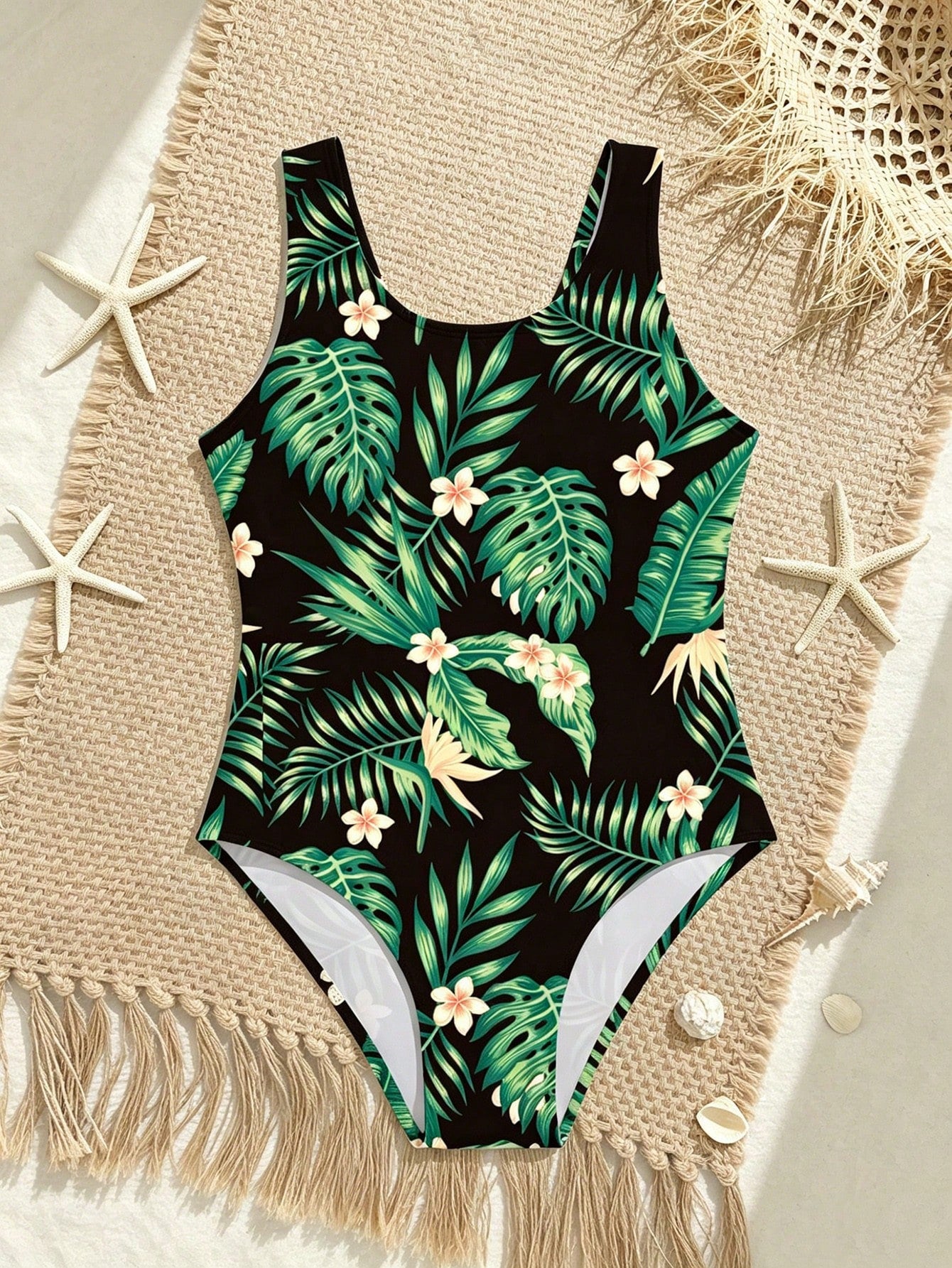 Tween girls one-piece swimsuit with a black base and multicolor tropical plant print featuring bamboo, palm leaves, plumeria flowers, scoop neck, and wide straps.