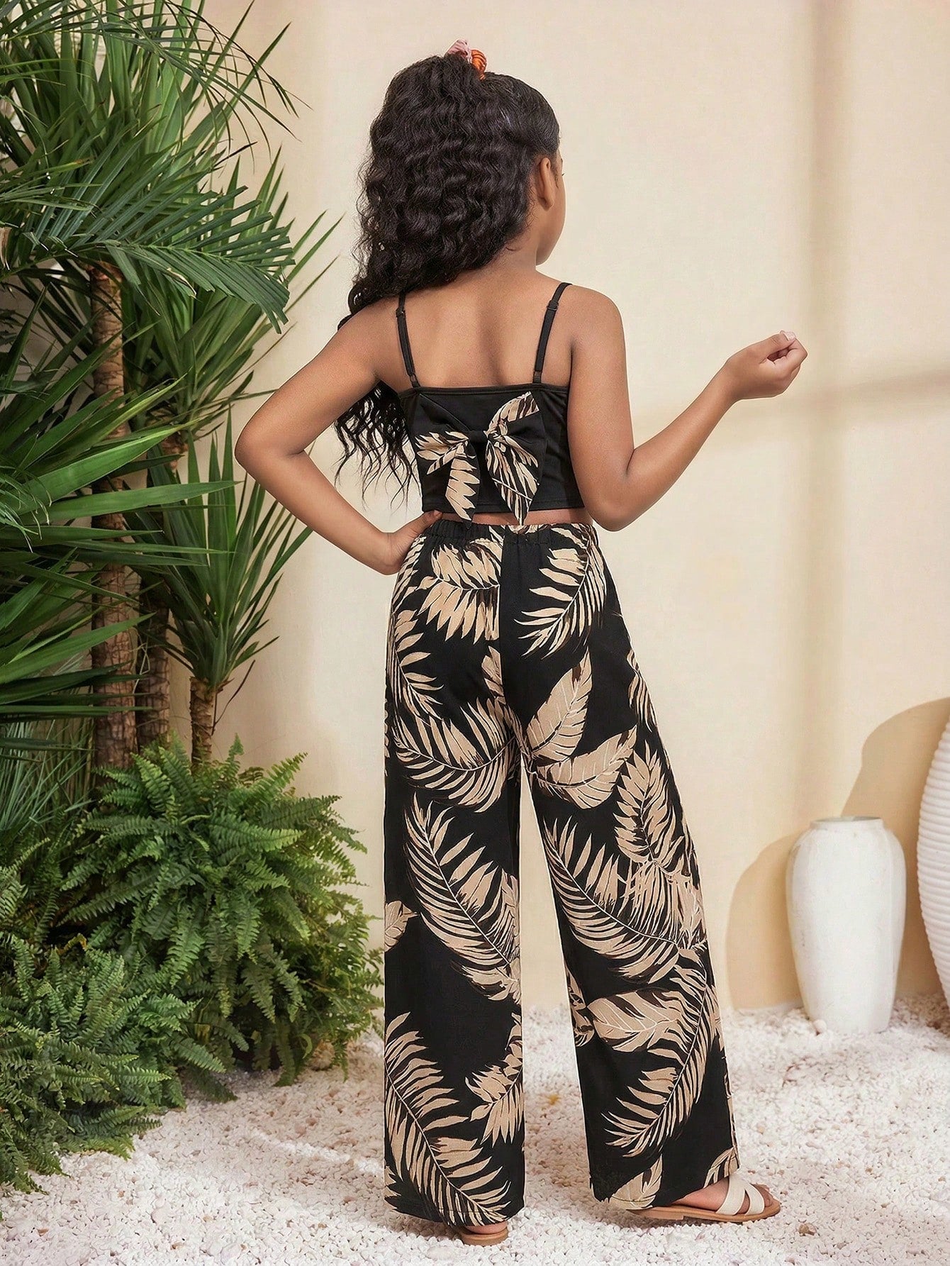 Tween girl wearing a black sleeveless spaghetti strap camisole top with 3D bow decor and matching leopard print wide leg long pants.