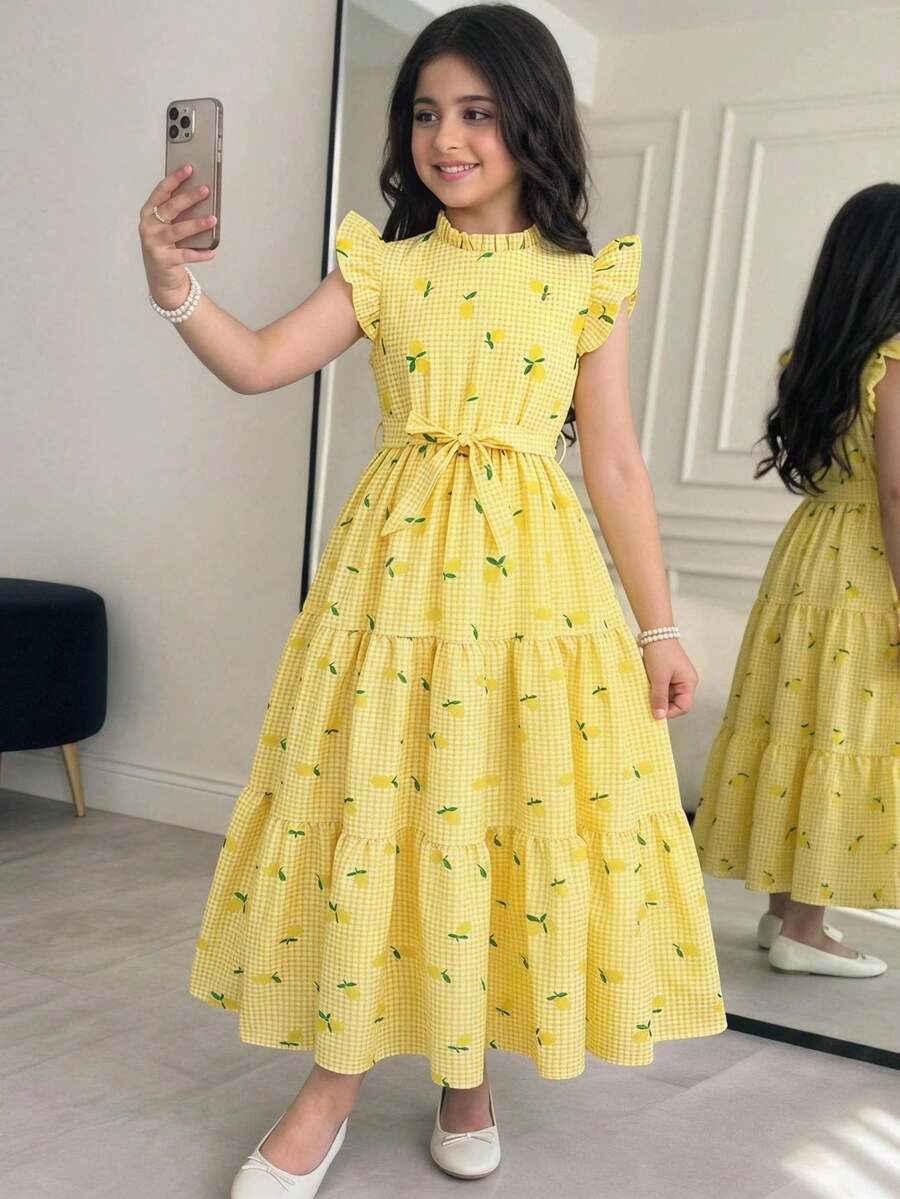 Tween girl wearing a pink long A-line dress featuring a strawberry print, stand collar, flounce short sleeves, and a belted waist with a ruffle hem.