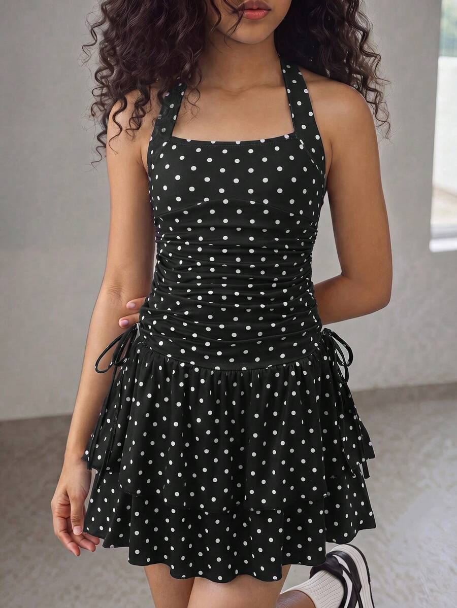 Tween girl wearing a black and white polka dot tiered cami dress with shoulder straps, a high waist, and a ruffle layered hem.