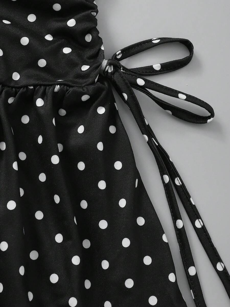 Tween girl wearing a black and white polka dot tiered cami dress with shoulder straps, a high waist, and a ruffle layered hem.
