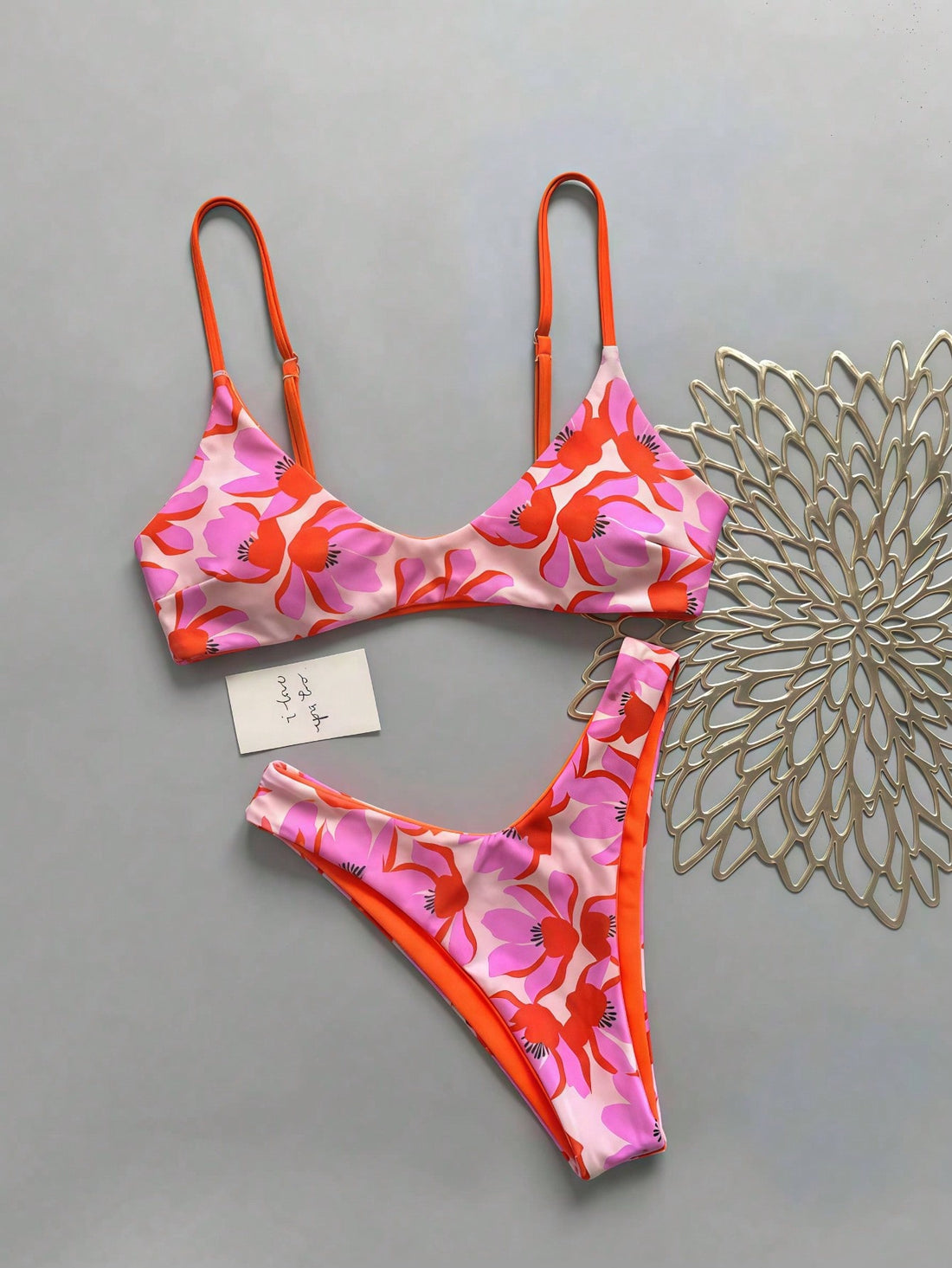 Women's pinkish orange floral print 2-piece bikini set featuring a backless wireless top with spaghetti straps and matching mid-waist bikini bottoms.