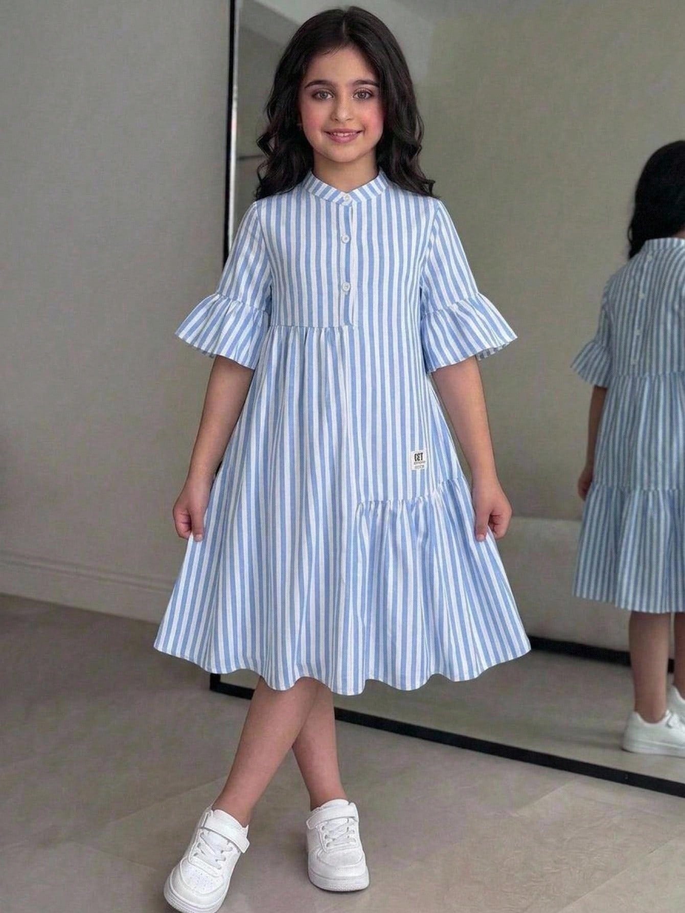 Tween girl wearing a black striped smock shirt dress with flounce half sleeves, a stand collar, and a ruffled hem.