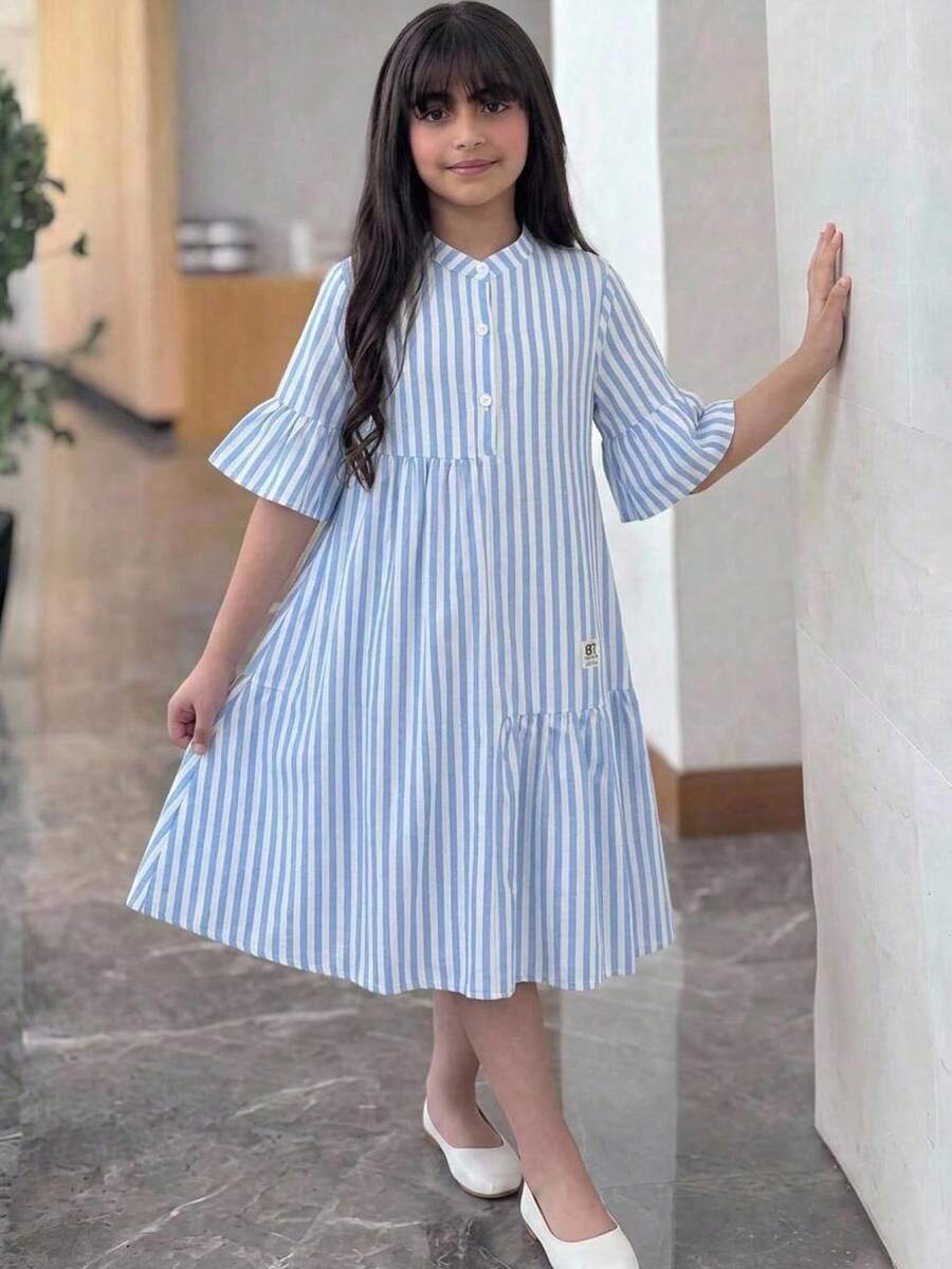 Tween girl wearing a black striped smock shirt dress with flounce half sleeves, a stand collar, and a ruffled hem.