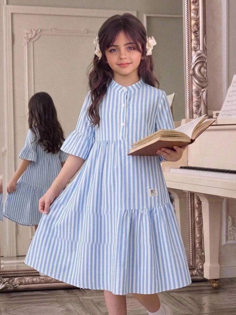 Tween girl wearing a black striped smock shirt dress with flounce half sleeves, a stand collar, and a ruffled hem.