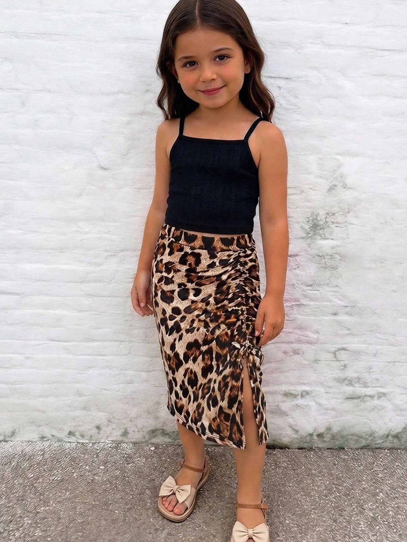 Tween girl wearing a 2-piece set featuring a black rib-knit spaghetti strap camisole top and a ruched leopard print midi skirt.