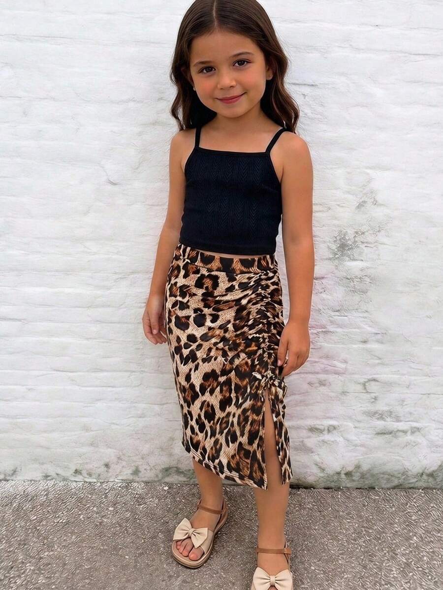Tween girl wearing a 2-piece set featuring a black rib-knit spaghetti strap camisole top and a ruched leopard print midi skirt.