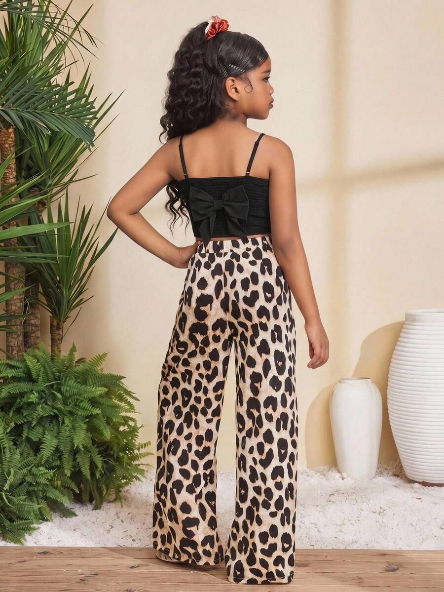 Tween girl wearing a black sleeveless spaghetti strap camisole top with 3D bow decor and matching leopard print wide leg long pants.